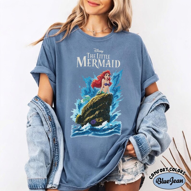 May include: A blue crew neck t-shirt featuring a graphic of Ariel from The Little Mermaid sitting on a rock surrounded by blue ocean waves. The text Disney The Little Mermaid is printed in white above the character. The shirt has short sleeves.