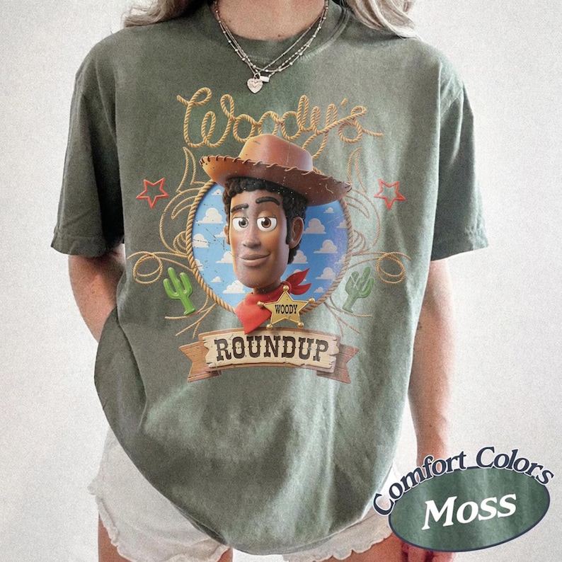 K&ouml;nnte beinhalten: Ein moosgr&uuml;nes Kurzarm-T-Shirt mit einer Grafik der Figur Woody aus Toy Story, die einen Cowboyhut und ein rotes Halstuch tr&auml;gt. Das Design zeigt einen blauen Himmel, Seilakzente, gr&uuml;ne Kakteen, rote Sterne und den Schriftzug Woody's Roundup.