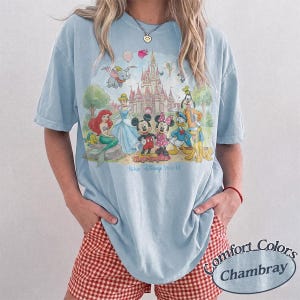 May include: A light blue short-sleeve t-shirt featuring a graphic print of various cartoon characters in front of a castle. The text "Magic Kingdom" and "Walt Disney World" is printed below the characters. The shirt has a crew neckline.