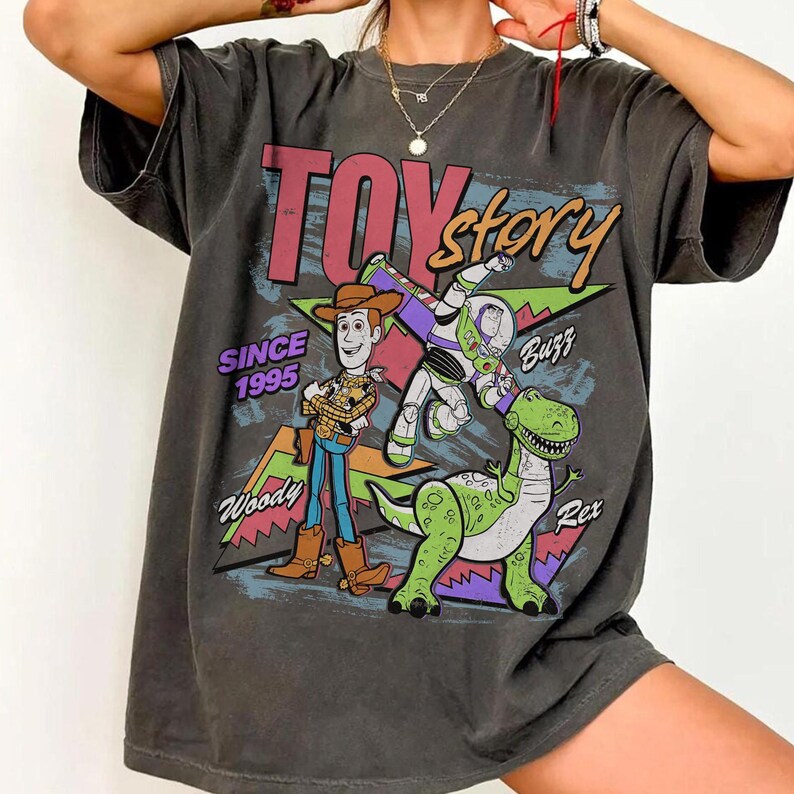 Pode incluir: T-shirt cinzento escuro com as palavras "Toy Story" e ilustra&ccedil;&otilde;es de Woody, Buzz Lightyear e Rex, o dinossauro. A camisa tamb&eacute;m inclui o texto "Since 1995" e os nomes dos personagens. O design usa vermelho, azul, verde e amarelo.