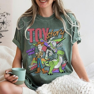 Pode incluir: T-shirt verde com o logotipo Toy Story e os personagens Woody, Buzz Lightyear e Rex. A camisa tem o texto "TOY STORY" e "SINCE 1995". O design inclui um gr&aacute;fico retr&ocirc; e colorido.
