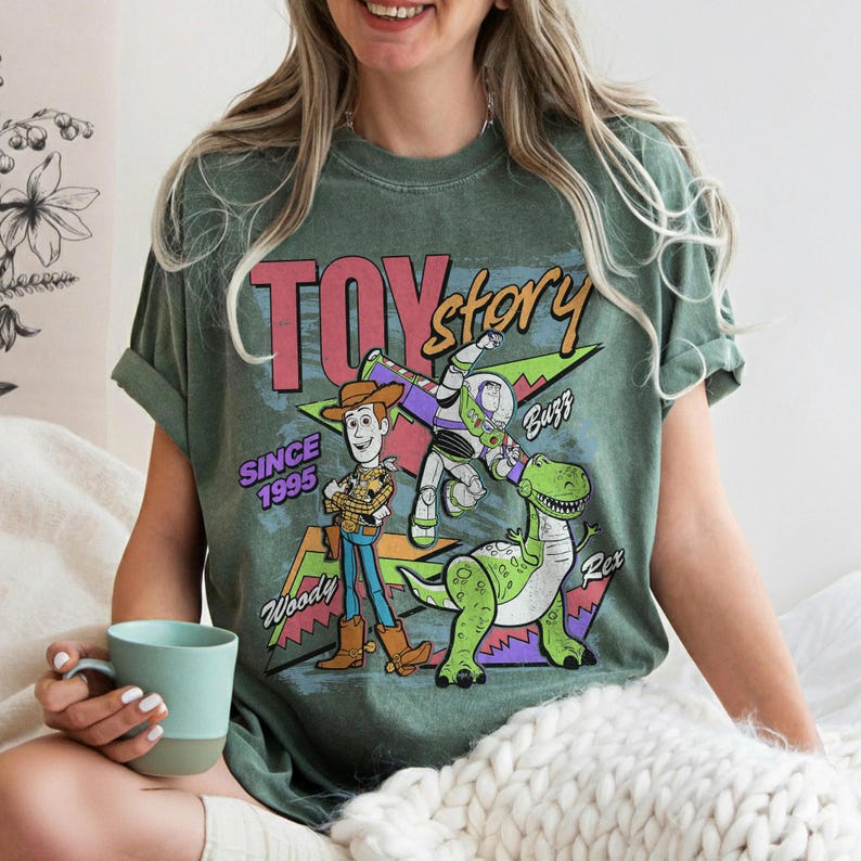 Pode incluir: T-shirt verde com o logotipo Toy Story e os personagens Woody, Buzz Lightyear e Rex. A camisa tem o texto "TOY STORY" e "SINCE 1995". O design inclui um gr&aacute;fico retr&ocirc; e colorido.