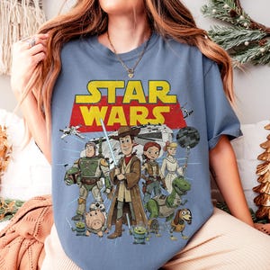 May include: A light blue t-shirt featuring a graphic of Star Wars characters, including Woody as Obi-Wan Kenobi. The Star Wars logo is in yellow and red. Other characters include Buzz Lightyear, Rex, and Bo Peep.