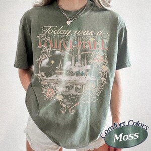 Puede incluir: Una camiseta de manga corta color verde musgo con un estampado gr&aacute;fico de un castillo, una mariposa y elementos florales. El texto dice Today was a FAIRY TALE y Can you feel the magic in the air? La camiseta tiene cuello redondo y corte holgado.