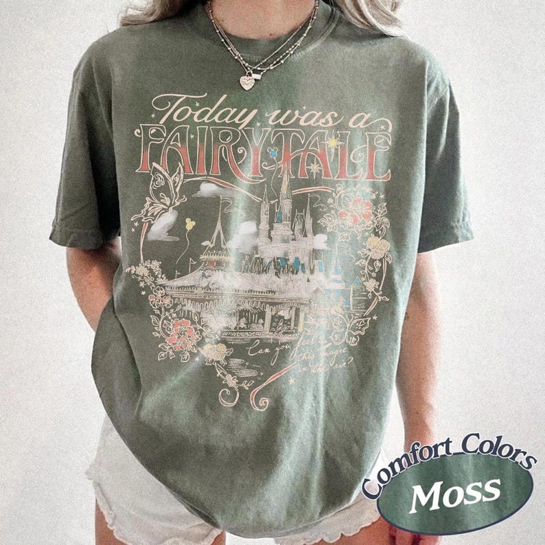 Puede incluir: Una camiseta de manga corta color verde musgo con un estampado gr&aacute;fico de un castillo, una mariposa y elementos florales. El texto dice Today was a FAIRY TALE y Can you feel the magic in the air? La camiseta tiene cuello redondo y corte holgado.