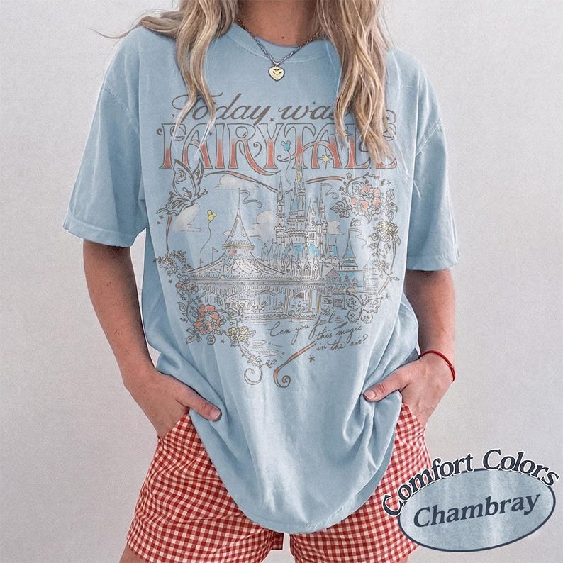 Puede incluir: Una camiseta de manga corta de color azul claro con un estampado gr&aacute;fico de un castillo, flores y una mariposa. El texto dice Today was a FAIRYTALE y Can you feel this magic in the air? La camiseta tiene un cuello redondo.