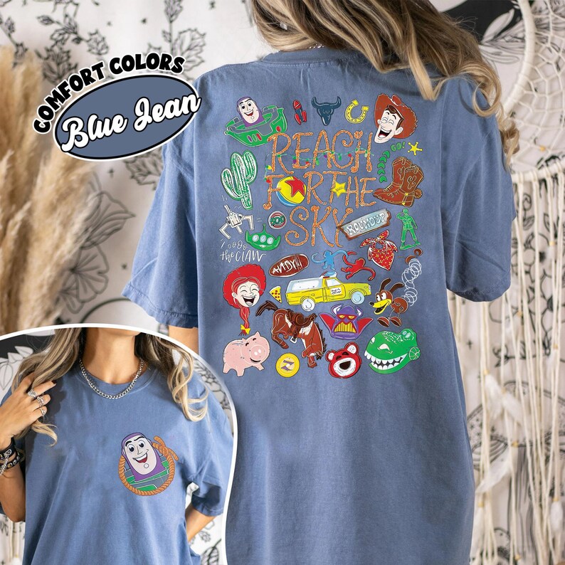 K&ouml;nnte beinhalten: Ein blaues T-Shirt mit einer kleinen Buzz Lightyear-Grafik auf der linken Brust. Die R&uuml;ckseite zeigt eine gro&szlig;e Collage aus Toy Story-Figuren und Symbolen mit dem Text Reach For The Sky und Comfort Colors Blue Jean.