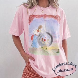 May include: A light pink short-sleeve t-shirt featuring a watercolor-style graphic of a person standing next to a tire swing. The illustration includes a landscape background and small figures inside the tire. The text Comfort Colors Blossom is visible.