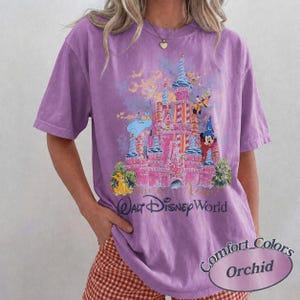 Może przedstawiać: Koszulka w kolorze orchidei z grafiką r&oacute;żowego zamku, postaciami Disneya i napisem "Walt Disney World". Koszulka ma kr&oacute;tkie rękawy i luźny kr&oacute;j. Widoczny jest r&oacute;wnież tekst "Comfort Colors" i "Orchid".