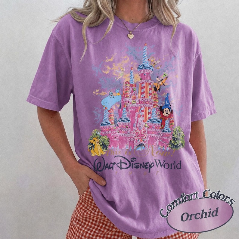 Może przedstawiać: Koszulka w kolorze orchidei z grafiką r&oacute;żowego zamku, postaciami Disneya i napisem "Walt Disney World". Koszulka ma kr&oacute;tkie rękawy i luźny kr&oacute;j. Widoczny jest r&oacute;wnież tekst "Comfort Colors" i "Orchid".