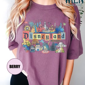 May include: Purple t-shirt with a vintage-style illustration of Disneyland Resort. The illustration includes iconic landmarks like Sleeping Beauty Castle, Space Mountain, and the Matterhorn.