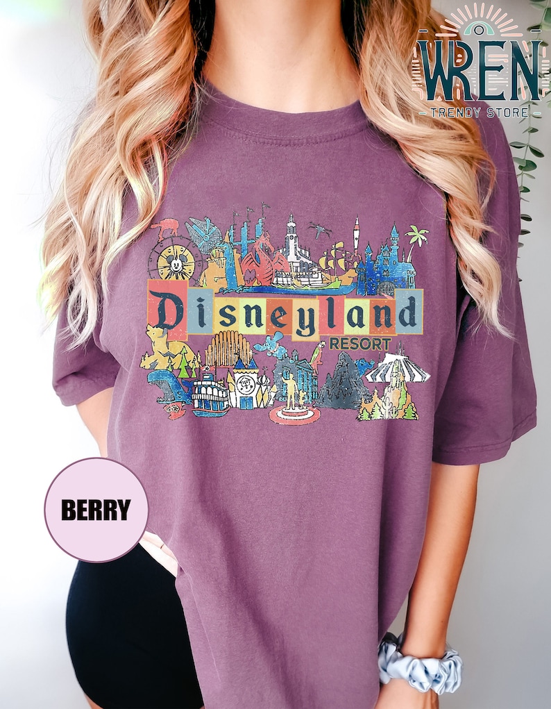 May include: Purple t-shirt with a vintage-style illustration of Disneyland Resort. The illustration includes iconic landmarks like Sleeping Beauty Castle, Space Mountain, and the Matterhorn.
