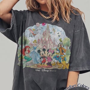 May include: A charcoal gray t-shirt featuring a graphic print of Disney characters including Mickey Mouse, Minnie Mouse, Donald Duck, Goofy, Pluto, Ariel, Cinderella, Dumbo, and Tinker Bell in front of a castle. Text reads Magic Kingdom Walt Disney World.