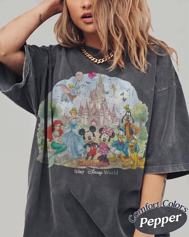 May include: A charcoal gray t-shirt featuring a graphic print of Disney characters including Mickey Mouse, Minnie Mouse, Donald Duck, Goofy, Pluto, Ariel, Cinderella, Dumbo, and Tinker Bell in front of a castle. Text reads Magic Kingdom Walt Disney World.