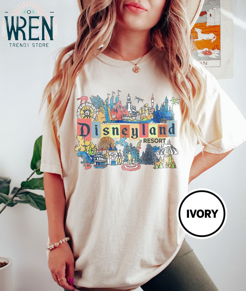 May include: A cream colored t-shirt with a colorful graphic of Disneyland Resort. The graphic includes iconic attractions like Sleeping Beauty's Castle, the Matterhorn, and the Haunted Mansion. The text "Disneyland Resort" is printed in red and yellow.