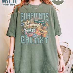 K&ouml;nnte beinhalten: Ein gr&uuml;nes T-Shirt mit einem grafischen Druck von Kassetten und Planeten. Der Text "Guardians of the Galaxy" ist auf dem T-Shirt gedruckt.