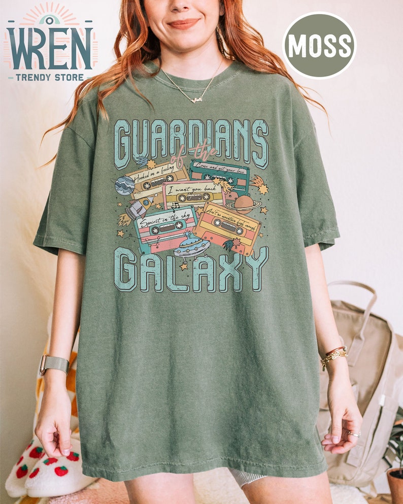 K&ouml;nnte beinhalten: Ein gr&uuml;nes T-Shirt mit einem grafischen Druck von Kassetten und Planeten. Der Text "Guardians of the Galaxy" ist auf dem T-Shirt gedruckt.