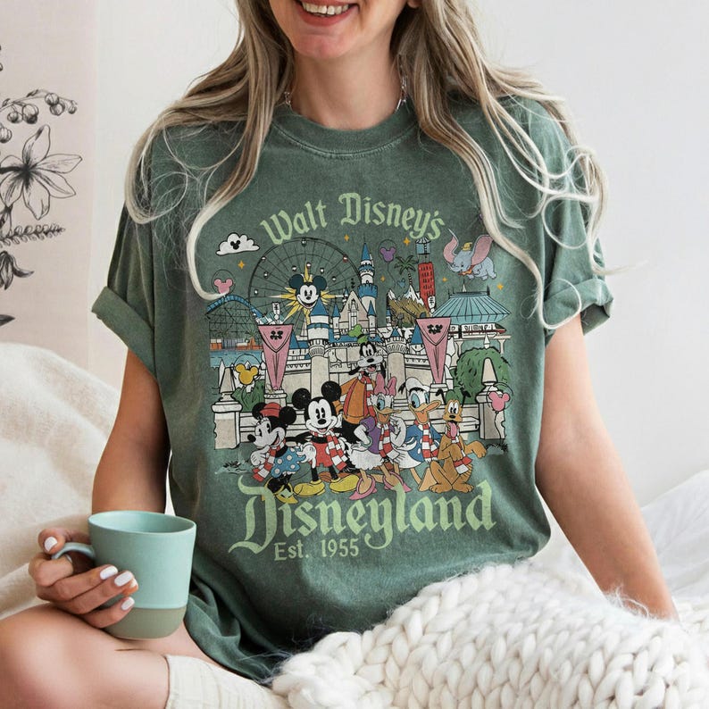 May include: Green t-shirt featuring a Disneyland graphic with Mickey Mouse, Minnie Mouse, Donald Duck, and other Disney characters. The shirt has the text "Walt Disney's Disneyland Est. 1955" in a vintage style.