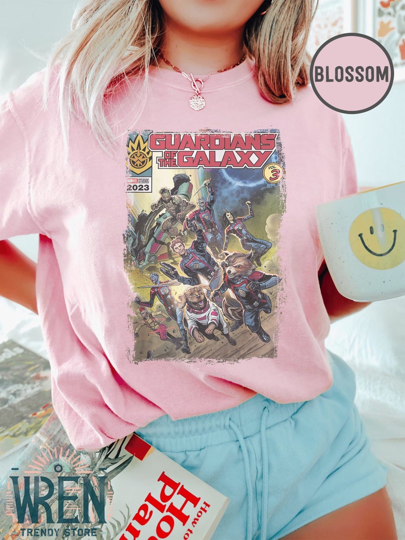 Pu&ograve; includere: Una maglietta rosa con un design di fumetto vintage con i Guardiani della Galassia. La copertina del fumetto ha il titolo "Guardiani della Galassia" e il numero "3" in un cerchio. L'anno "2023" &egrave; stampato sulla copertina.