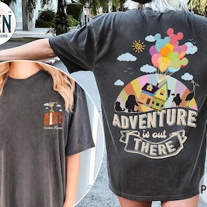 May include: Dark grey t-shirt with a colourful graphic on the back featuring a house lifted by balloons, the text "Adventure is out there", and a small graphic on the front. The shirt is made of a soft material.