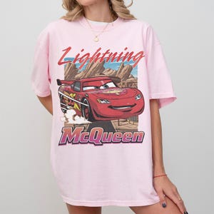 May include: Light pink t-shirt featuring a graphic of Lightning McQueen from the Cars movie. The design includes the text "Lightning McQueen" in red and purple, with a cartoon image of the red race car.