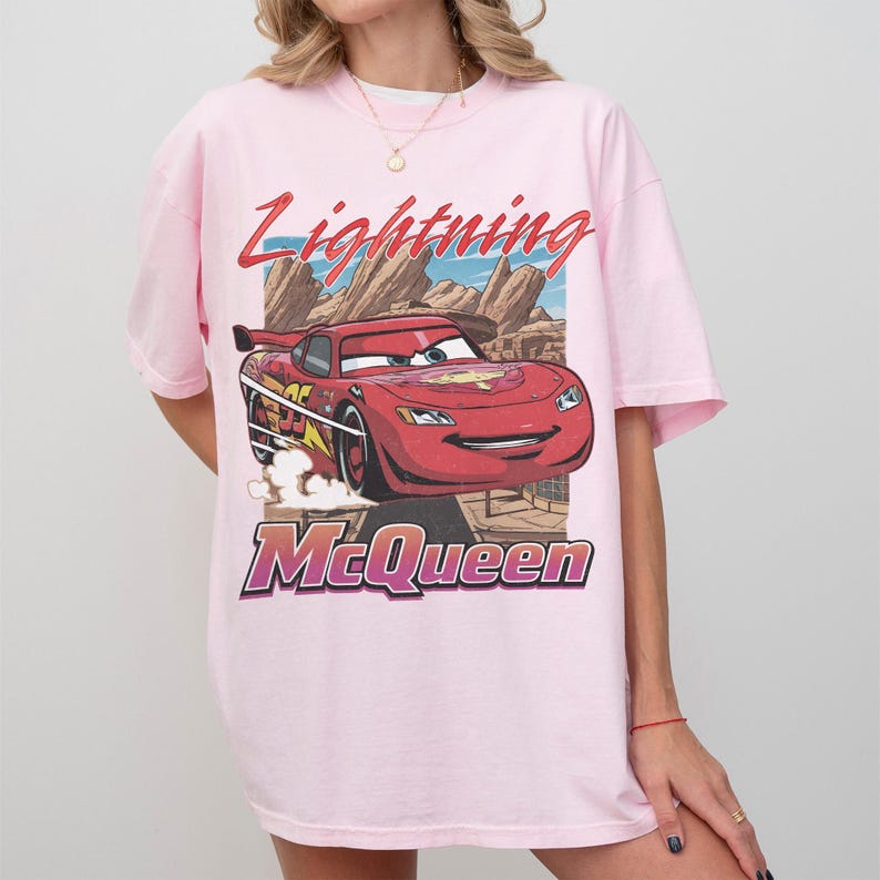 May include: Light pink t-shirt featuring a graphic of Lightning McQueen from the Cars movie. The design includes the text "Lightning McQueen" in red and purple, with a cartoon image of the red race car.