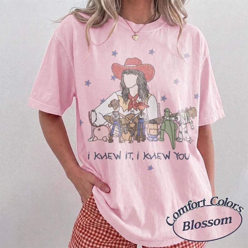 May include: A light pink short-sleeve t-shirt featuring a graphic illustration of a cowgirl and various toy characters. The text "I KNEW IT, I KNEW YOU" is printed below the graphic, surrounded by small blue stars on the front of the garment.