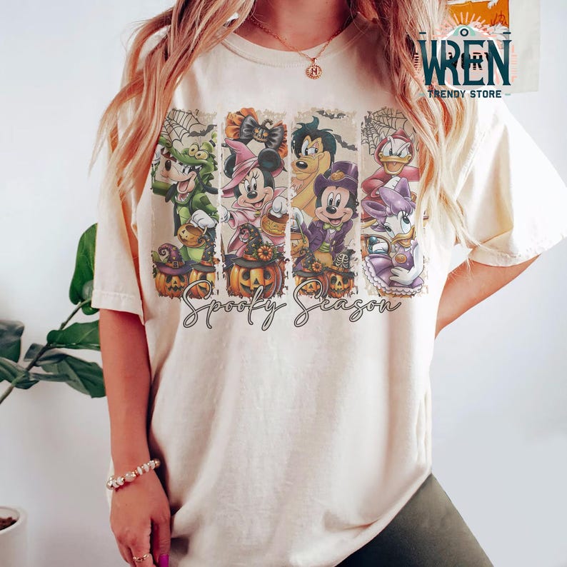 May include: Cream-colored t-shirt featuring a Halloween-themed graphic with cartoon characters. The design includes images of Mickey Mouse, Minnie Mouse, Donald Duck, and Goofy in Halloween costumes, along with the words "Spooky Season".