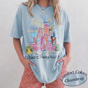 Może przedstawiać: Jasnoniebieska koszulka z r&oacute;żowym zamkiem, postaciami Disneya i napisem "This is Now! Walt Disney World". Wykonana z tkaniny Comfort Colors Chambray.