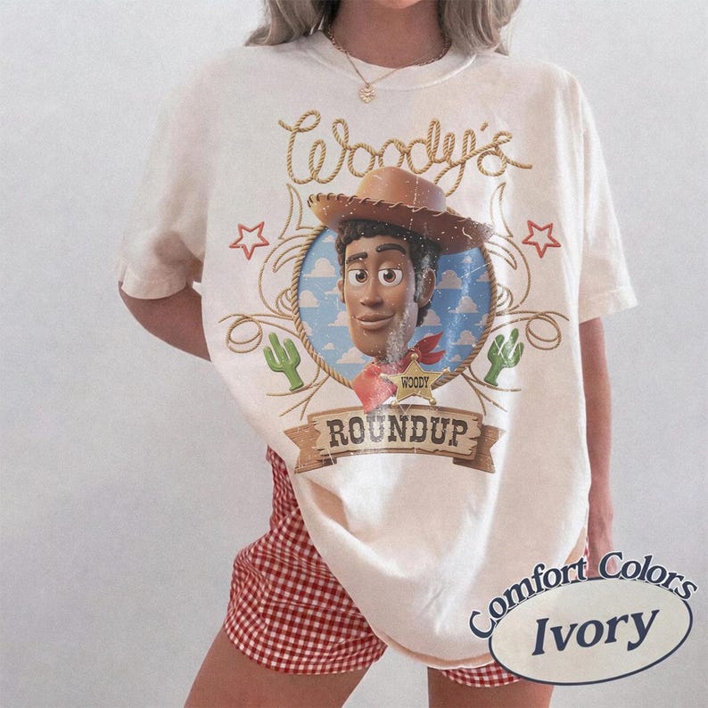 K&ouml;nnte beinhalten: Ein elfenbeinfarbenes T-Shirt mit einer Grafik der Figur Woody aus Toy Story in einem kreisf&ouml;rmigen Rahmen. Das Design enth&auml;lt den Text Woody's und ROUNDUP sowie Seilakzente, Sterne und Kaktusillustrationen im Used-Look.