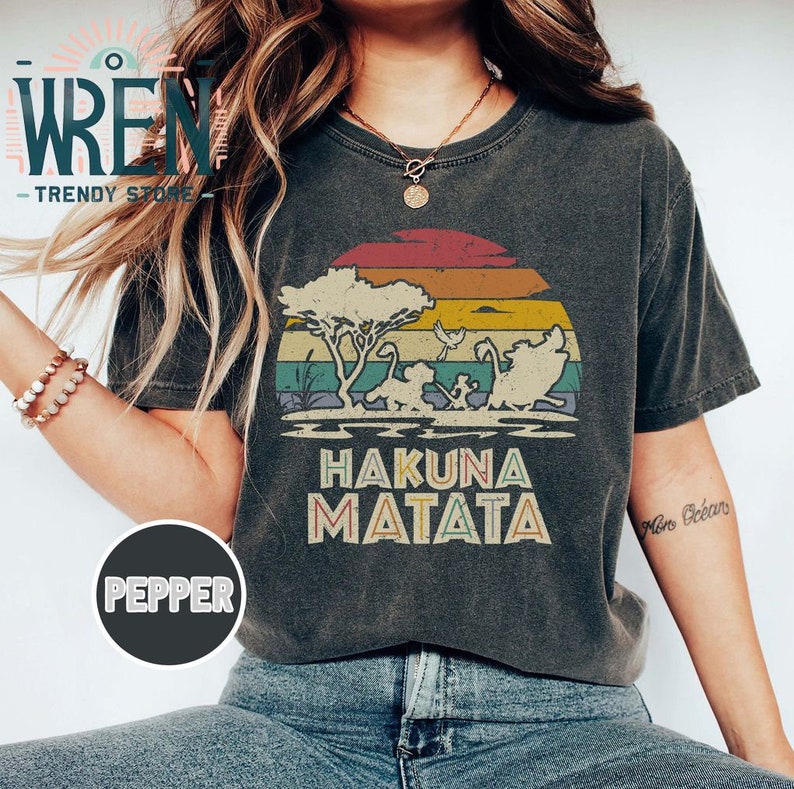 May include: A dark gray t-shirt with a vintage-style sunset graphic featuring a silhouette of the Lion King characters. The text "Hakuna Matata" is printed below the graphic in a retro font.