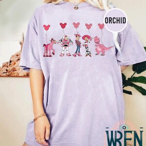 Może przedstawiać: Jasno-fioletowy t-shirt z grafiką postaci z Toy Story trzymających r&oacute;żowe balony w kształcie serca. Postacie to Woody, Buzz Astral, Jessie i Rex. Tekst "ORCHID" znajduje się w prawym g&oacute;rnym rogu obrazu.