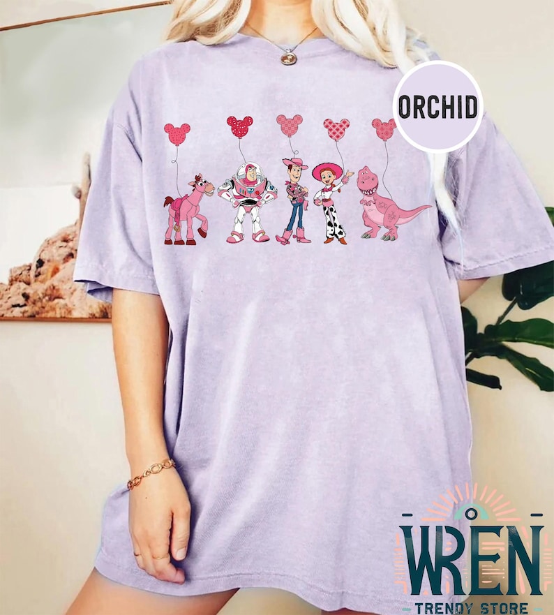 Może przedstawiać: Jasno-fioletowy t-shirt z grafiką postaci z Toy Story trzymających r&oacute;żowe balony w kształcie serca. Postacie to Woody, Buzz Astral, Jessie i Rex. Tekst "ORCHID" znajduje się w prawym g&oacute;rnym rogu obrazu.