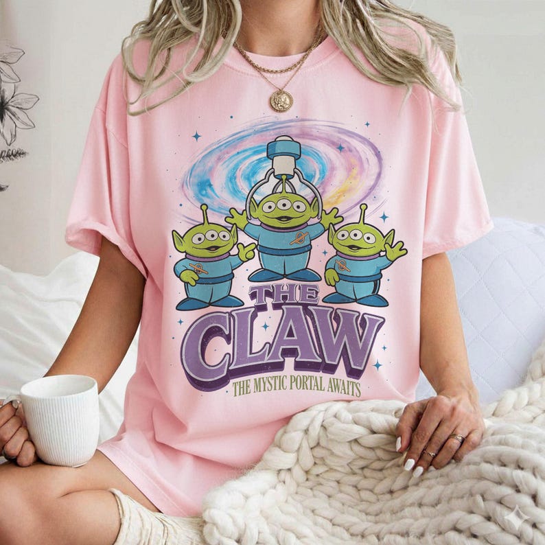 以下が含まれることがあります： ピンク色のTシャツ。青いスーツを着た3人の緑色のエイリアンのデザインで、紫と白で「THE CLAW」の文字が描かれています。カラフルな銀河の背景と「THE MYSTIC PORTAL AWAITS」のフレーズも含まれています。