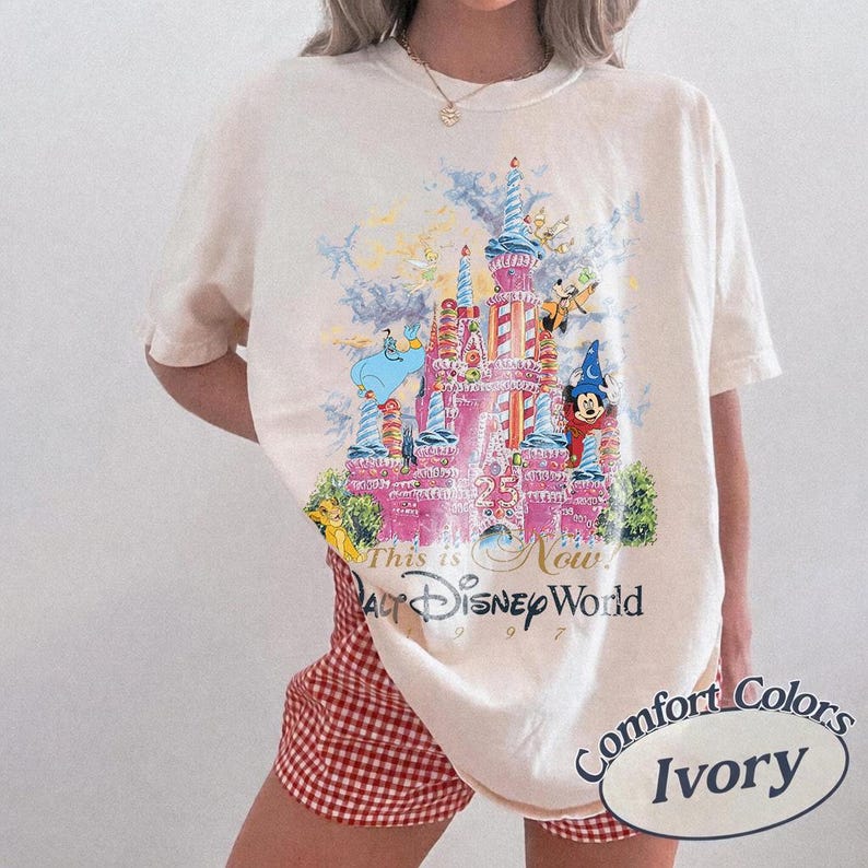 May include: Ivory-colored t-shirt featuring a colorful graphic of Cinderella's castle with Disney characters. The text "This is Now! Walt Disney World 1997" is printed below the castle. The shirt is a Comfort Colors brand.