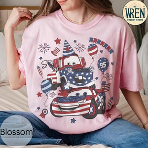 Puede incluir: Camiseta rosa claro con un dise&ntilde;o de dos coches de dibujos animados decorados con patrones de la bandera estadounidense, fuegos artificiales y globos. El dise&ntilde;o incluye el n&uacute;mero 95 y elementos patri&oacute;ticos.