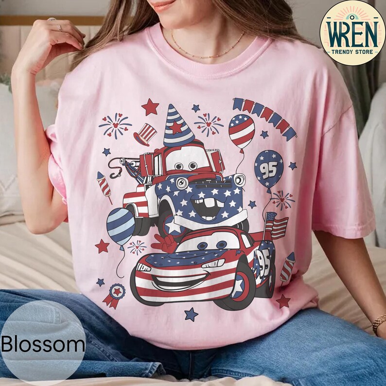 Puede incluir: Camiseta rosa claro con un dise&ntilde;o de dos coches de dibujos animados decorados con patrones de la bandera estadounidense, fuegos artificiales y globos. El dise&ntilde;o incluye el n&uacute;mero 95 y elementos patri&oacute;ticos.