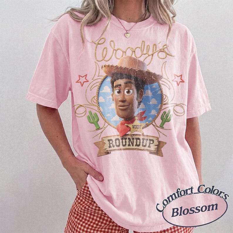 K&ouml;nnte beinhalten: Ein hellrosa T-Shirt mit kurzen &Auml;rmeln und einer Grafik der Figur Woody aus Toy Story in einem kreisf&ouml;rmigen Rahmen. Das Design enth&auml;lt den Text Woody's ROUNDUP, eine Seilumrandung, Sterne und Kaktussymbole. Das Shirt hat einen Rundhalsausschnitt.