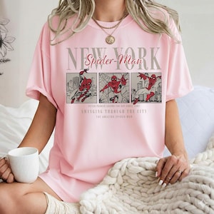 Pu&ograve; includere: T-shirt rosa chiaro con grafica di Spider-Man. Il design include le parole "New York" e "Spider-Man" sopra tre pannelli che raffigurano Spider-Man in azione. Il testo sottostante recita "Peter Parker Saving the Day Since 1982" e "Swinging Through the City."