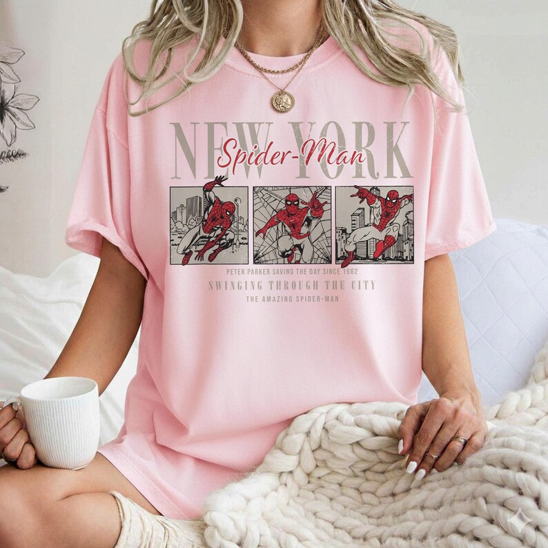 Pu&ograve; includere: T-shirt rosa chiaro con grafica di Spider-Man. Il design include le parole "New York" e "Spider-Man" sopra tre pannelli che raffigurano Spider-Man in azione. Il testo sottostante recita "Peter Parker Saving the Day Since 1982" e "Swinging Through the City."