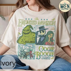 以下が含まれることがあります： 「The Haunted Mansion」と「Oogie Boogie Bash」のキャラクターが描かれたアイボリー色のTシャツ。デザインにはジャック・スケリントン、サリー、ウギー・ブギーが含まれています。「The Haunted Mansion」と「Oogie Boogie Bash」の文字も表示されています。
