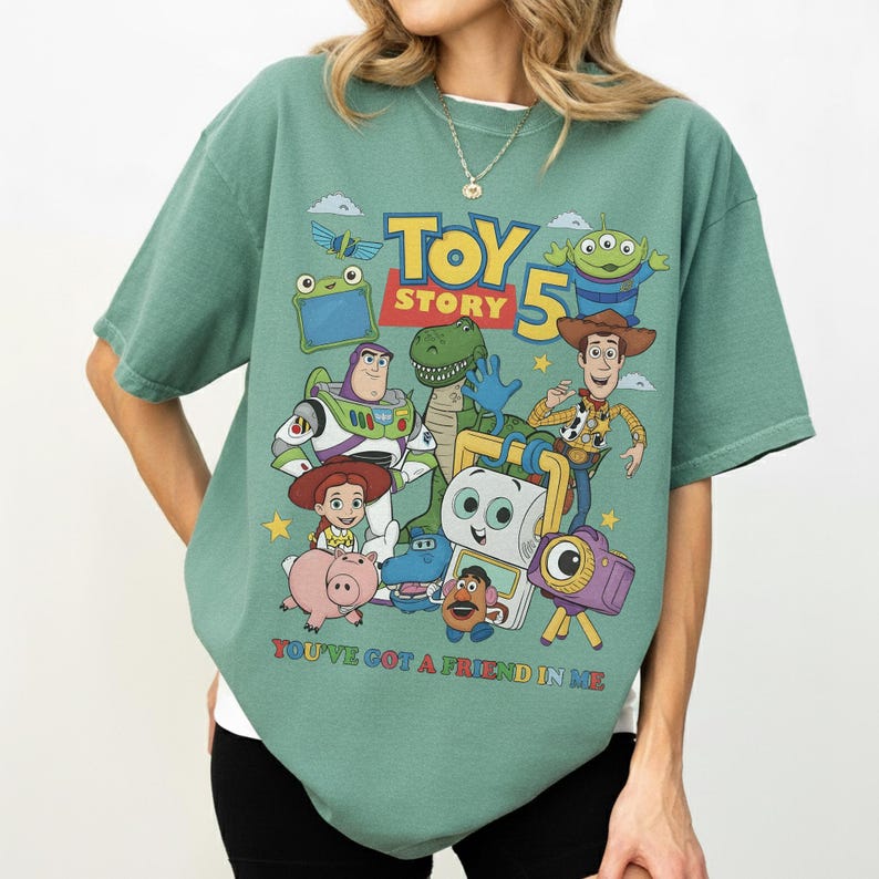 May include: A green short-sleeve t-shirt featuring a colorful graphic of characters from the animated film "Toy Story." The design includes Woody, Buzz Lightyear, and other characters, with the text "Toy Story 5" and "You've Got a Friend in Me."