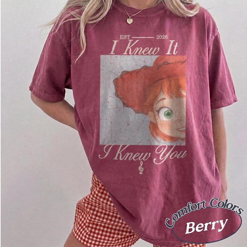 May include: A berry-colored short-sleeve t-shirt featuring a graphic of a red-haired character in a hat. The text reads EST 2026 I Knew It I Knew You with a treble clef symbol. The shirt has a distressed, vintage-style print on the front center.