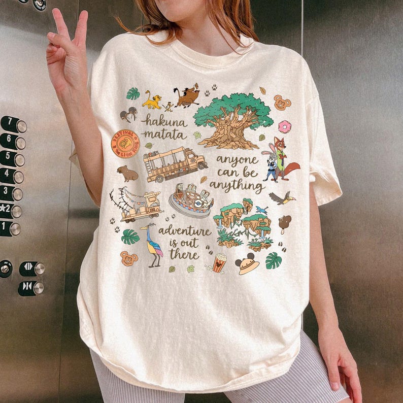May include: Cream-colored t-shirt featuring colorful cartoon illustrations and text. The shirt includes phrases like "Hakuna Matata" and "Adventure is out there." Various characters and icons are depicted, including a tree, animals, and a safari vehicle.