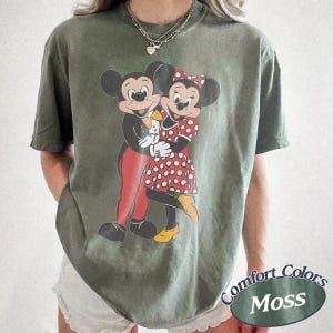 May include: A moss green short-sleeve t-shirt featuring a graphic print of Mickey and Minnie Mouse embracing. Mickey wears red pants and a yellow bowtie, while Minnie wears a red and white polka dot dress with yellow shoes. The shirt has a crew neck.