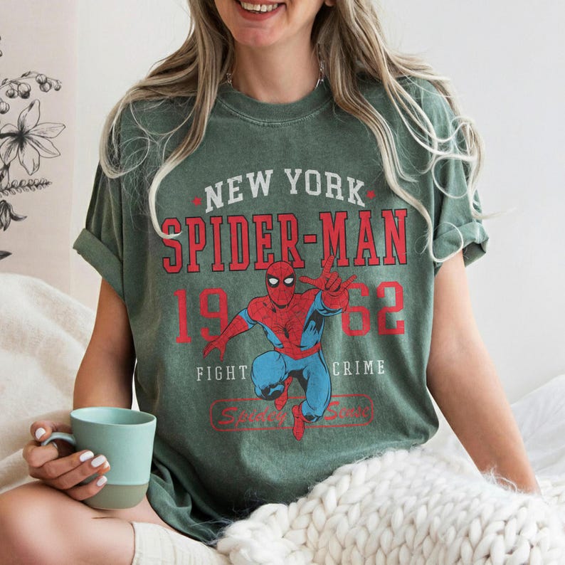 Op de afbeelding: Groen t-shirt met een Spider-Man afbeelding in rood en blauw, met de tekst "NEW YORK SPIDER-MAN 1962 FIGHT CRIME". Het shirt heeft opgerolde mouwen en een vintage uitstraling.