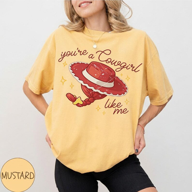 May include: A mustard yellow short-sleeve t-shirt featuring a graphic of a red cowgirl hat with a braided ponytail. The text reads you're a Cowgirl like me, accompanied by small yellow star icons. The design has a distressed, vintage-inspired texture.