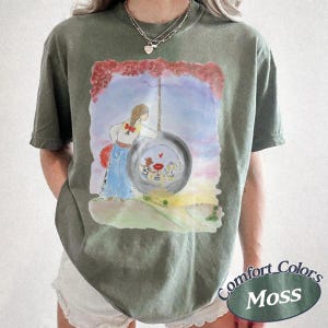May include: An olive green short-sleeve t-shirt featuring a watercolor-style graphic of a person standing next to a tire swing. The illustration includes a red-roofed house inside the tire, a red sky, and a grassy landscape. The shirt has a crew neck.