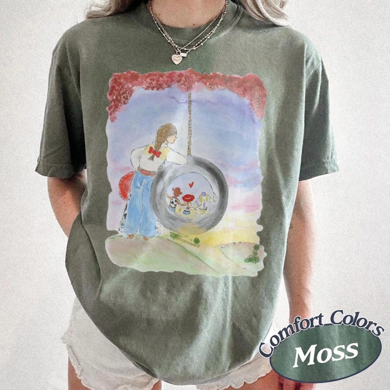 May include: An olive green short-sleeve t-shirt featuring a watercolor-style graphic of a person standing next to a tire swing. The illustration includes a red-roofed house inside the tire, a red sky, and a grassy landscape. The shirt has a crew neck.