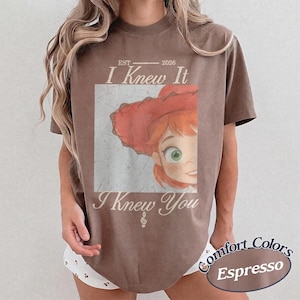 May include: A brown short-sleeve t-shirt featuring a graphic of a cartoon character with red hair and a red hat. The text reads EST 2026 I Knew It I Knew You with a small musical note symbol. The design has a distressed, vintage-style appearance.